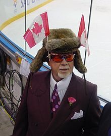  Photographie couleur de Don Cherry en costume-cravate dans les tribunes d'une patinoire. Il porte une chapka surmontée de deux drapeaux canadiens.