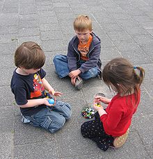 Trois enfants âgés d'environ six ans sont groupés assis sur le sol, un garçon et une fille agenouillés, l'autre garçon assis en tailleur. Les deux enfants agenouillés tiennent des billes ; il y a d'autres billes dans un sac sur le sol. Les trois enfants les regardent.