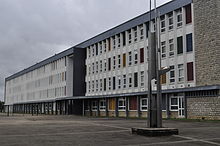 Un long bâtiment blanc aux panneaux colorés s'étend sur la droite de l'image. Au premier plan, on voit une sculpture moderne en fer.