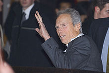 Photographie de Clint Eastwood saluant le public après une conférence de presse. Il est vêtu d'un costume gris foncé