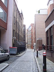 La photo montre une allée pavée de pierres taillées et qui est entourée d'immeubles.