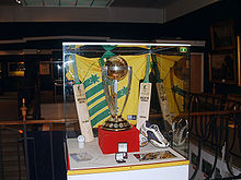 Photographie représentant le trophée de la Coupe du monde en vitrine devant le maillot utilisé par l'Australie en 1999.