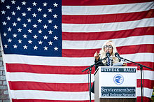 Une femme aux cheveux blonds portant d'imposantes lunettes parlant dans un microphone. Derrière elle se trouve un drapeau des États-Unis recouvrant un mur de brique.