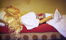 Un mannequin féminin artificiel est allongé sur un lit devant un mur à l’aspect ancien dont le fin matelas est rouge, tête vers la gauche. Habillée d’une robe blanche et d’une perruque de longs cheveux blonds, sa main droite tenant un mouchoir blanc est surélevée.