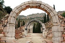 La photographie en couleur représente une vue de la chartreuse d'Escaladei. A travers deux portes voutées dont le mur est écroulé, apparait au fond l'entrée de style baroque de la chartreuse, au bout d'une allée bordée de cyprès. En arrière-plan, la sierra de montsant surplombe le site.