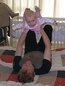 Un jeune père allongé sur le dos sur un édredon sur le sol. Il tient sa fille bébé au dessus de lui avec ses bras tendus et ses mains autour de sa cage thoracique. Le bébé a ses bras et ses jambes tendus et arque son dos, il sourit à l'appareil photo.