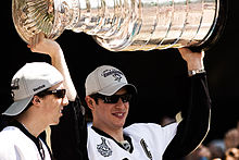 Photo de Sidney Crosby portant la Coupe Stanley au-dessus de sa tête à côté de Marc-André Fleury ; tous deux portent des lunettes de soleil et une casquette de champion.