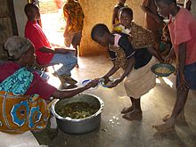 Dans une case, des enfants présentent leur assiette à une femme assise près d’un récipient métallique cabossé, posé à même le sol, et rempli d’une sorte de purée de légumes verts et orange.
