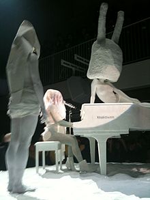 Photographie de Lady Gaga assise sur un banc, vêtue d'une robe traditionnelle de mariage japonais, jouant du piano tout en chantant.