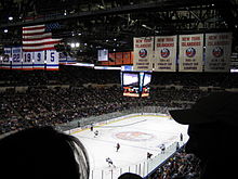 Photo de l'intérieur du Nassau Veterans Memorial Coliseum lors d'un match de hockey dont on aperçoit les joueurs en fond. On voit 4 bannières de champion de la coupe Stanley des Islanders de New York et 5 bannières de maillots retirés de ces mêmes Islanders.