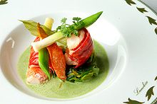 Au centre d'une assiette blanche, décorée de feuillages et marquée du nom Lameloise sur le bord, des morceaux de homard, surmontés de fragments de petits poireaux et de persil plat, trônent dans une sauce mousseuse verte.