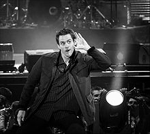 Photographie en noir et blanc de Jim Carrey en plein spectacle, il porte la main à son oreille comme pour mieux entendre