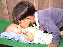 Sur la droite un jeune garçon d'apparence asiatique, les cheveux coupés en bol, se penche au dessus d'un bébé allongé sur le dos à gauche de l'image. Le garçon et le bébé se touchent par le nez. Le bébé regarde le garçon avec une expression d'intense intérêt.