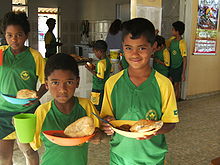 Groupe d’enfants en uniforme vert et jaune qui reçoivent au comptoir d’une cantine scolaire, une assiette avec un peu de nourriture et un énorme morceau de pain.