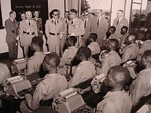photo sépia. Le roi en uniforme militaire, entouré d’officiels, fait face à un groupe d’élèves dactylographes.