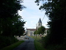Une petite route bordée d'arbres avec au fond une église de pierre couverte de tuiles au clocher conique ardoisé. Un panneau de limitation de vitesse à 30km/h à droite.