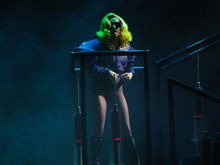 Photographie de Lady Gaga debout sur une plateforme, vêtue d'une veste mauve et d'une paire de lunettes de la même couleur.
