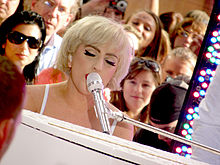 Une femme aux cheveux blonds courts, qui a les yeux fermés, jouant des notes sur un piano blanc tout en chantant. Derrière elle se trouvent plusieurs personnes l'observant.