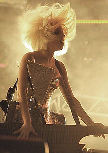 Photographie de Lady Gaga, vêtue d'une robe argentée, chantant sur scène tout en jouant du synthétiseur.