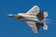 Photographie d’un F-22 effectuant un virage en postcombustion dans un ciel parfaitement bleu.