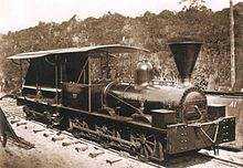 Une vieille photo montrant une locomotive d'un noir brillant avec une cabine aux côtés ouverts et une grande cheminée en forme d'entonnoir