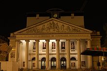 Photographie de nuit de la façade éclairée du théâtre : le péristyle aux huit colonnes surmontées du fronton sculpté précédant l'entrée du grand hall.