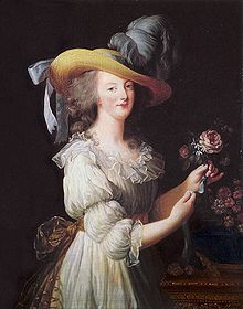 Portrait de trois-quarts d'une femme portant un grand chapeau de paille avec des plumes d'autruche bleu-gris, et une robe couleur crème avec un noeud doré à la ceinture dans le dos. Elle arrange un bouquet de fleurs roses.