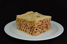 Photographie couleur montrant une assiette blanche sur fond noir garnie. Sous l'abaisse de pâte, les macaronis sont soudés par la farce viande-tomate-fromage et la découpe est parfaitement cubique.