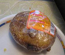 Posée sur une assiette, une grosse pomme de terre emballée d’un plastique transparent présente son étiquette orange avec les mentions de marque et temps de cuisson, et un code barre blanc et noir.