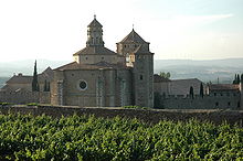 La photographie couleur représente une vigne en période végétative devant un mur d'enceinte. En arrière-plan apparaîtl'église du monastère de Poblet.