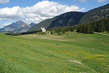 La photo couleur présente un alpage fleuri et valonné au premier plan. En arrière plan, un monument dédié à la Résistance sur le plateau des Glières se découpe face aux versants boisés et rocheux du fond de l'image.