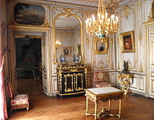Pièce comportant des boiseries blanches et dorées, parquet au sol et lustre doré, une porte à gauche surmonté d'un tableau, un bahut de couleur noire surmonté d'un miroir et plusieurs portraits peints aux murs