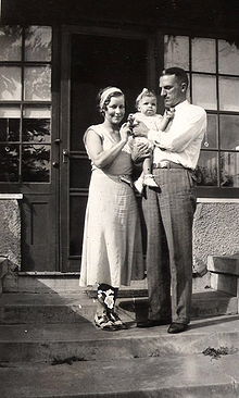 Un couple se tient debout devant une maison. L'homme, dans la trentaine, est habillé d'un pantalon de flanelle grise et d'une chemise blanche, tient une petite fille dans ses bras et la regarde. La femme, vêtue d'une robe et de chaussures des années 1930, se tient près d'eux, touchant le bébé et souriant à l'appareil photo. Le bébé est habillé d'une robe blanche, de chaussures blanches, et porte un ruban blanc dans les cheveux.