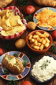 Parmi quelques tubercules de couleurs différentes, cinq assiettes posées sur une table présentent des chips, une pomme de terre de robe des champs, de la purée, des croquettes et une galette de pomme de terre.