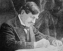 Paul Bourget vers 1893, assis à une table en train d'écrire, porte plume dans la main droite et bras gauche appuyé sur le bureau, visage penché et regard concentré sur son manuscrit.