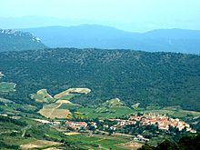 La photographie couleur présente une vue aérienne d'un vallon des Corbières. Le village est au fond, entouré de vignes sur les coteaux les moins pentus. Le sommet est boisé. En arrière-plan, le mont Tauch se devine en silhouette bleutée.