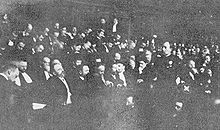 Photo du procès de Zola en 1898. Zola est de profil, sur la gauche de la photographie, entouré de différents protagonistes présents lors du procès. Il porte moustache, barbe et lunettes ; la photographie est trouble.