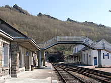 Le bâtiment voyageurs, la passerelle, les voies et quais, avec en arrière plan, les roches des Quatre Fils Aymon et l'entrée du tunnel de Château-Regnault