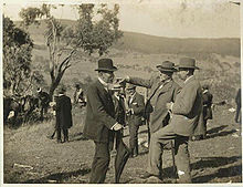 Trois hommes barbus d'âge moyen avec costume et chapeau sont debout dans un champ vallonné avec un arbre à proximité