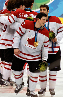Photographie couleur de Crosby dans le maillot du Canada avec la médaille d'or des Jeux olympiques autour du coup devant trois de ses coéquipiers.