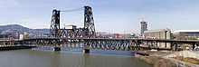 Le pont Steel Bridge est marqué par deux tours de fer qui maintiennent le pont, lui-même en fer. La rivière Willamette passe dessous. Des deux côtés du pont, quelques bâtiments de Portland apparaissent.