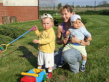 Une jeune mère est agenouillée dans un jardin avec ses deux enfants. Un bébé est assis à califourchon sur ses genoux, tourné vers l'avant et ne regarde pas l'appareil photo. Un jeune enfant se tient debout légèrement en avant de sa mère, tient une bêche et fronce les sourcils à l'appareil photo.