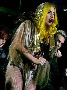 Photographie de Lady Gaga debout, vêtue d'une veste dorée accesoirisée d'un pelage et d'un justaucorps de cuir, chantant.