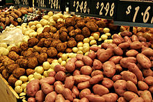 Gros plan sur un étal où les pommes de terre vendues en vrac sont rangées par variétés et couleurs alternées : roses, jaune paille, brun, jaune paille, etc. ; à l’arrière des ardoises en indiquent le prix en dollars par kg.