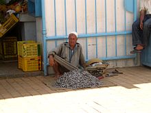 Vendeur de poisson assis sur le trottoir et appuyé contre le mur ; la marchandise est déposée à ses pieds.