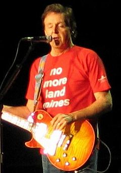 McCartney, jouant sur une guitare électrique orange et portant un T-shirt rouge où est écrit No More Land Mines.