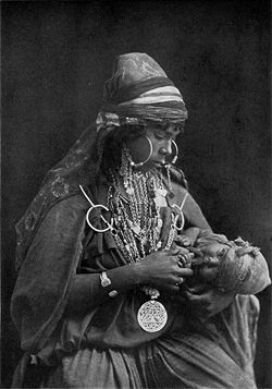 Photo présentant une mère bédouine tenant son enfant dans ses bras, son regard est fixé sur l'enfant qui semble jouer avec une partie du vêtement de sa mère tout en lui tenant un doigt