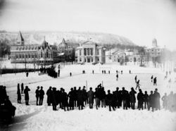 Photo noir et blanc d'un match de hockey joué en extérieur devant une ville enneigée.
