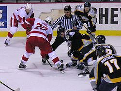Photo d'un match de hockey où un joueur des Red WIngs dispute le palet à une joueur de l'équipe adverse lors de la mise en jeu effectuée par un arbitre, le tout sous les yeux de leurs coéquipiers.