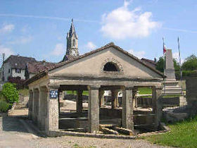 Photo du lavoir de Fêche-l'Église avec plus au fond à droite le monument aux morts et derrière le lavoir on aperçoit le clocher de l'église
